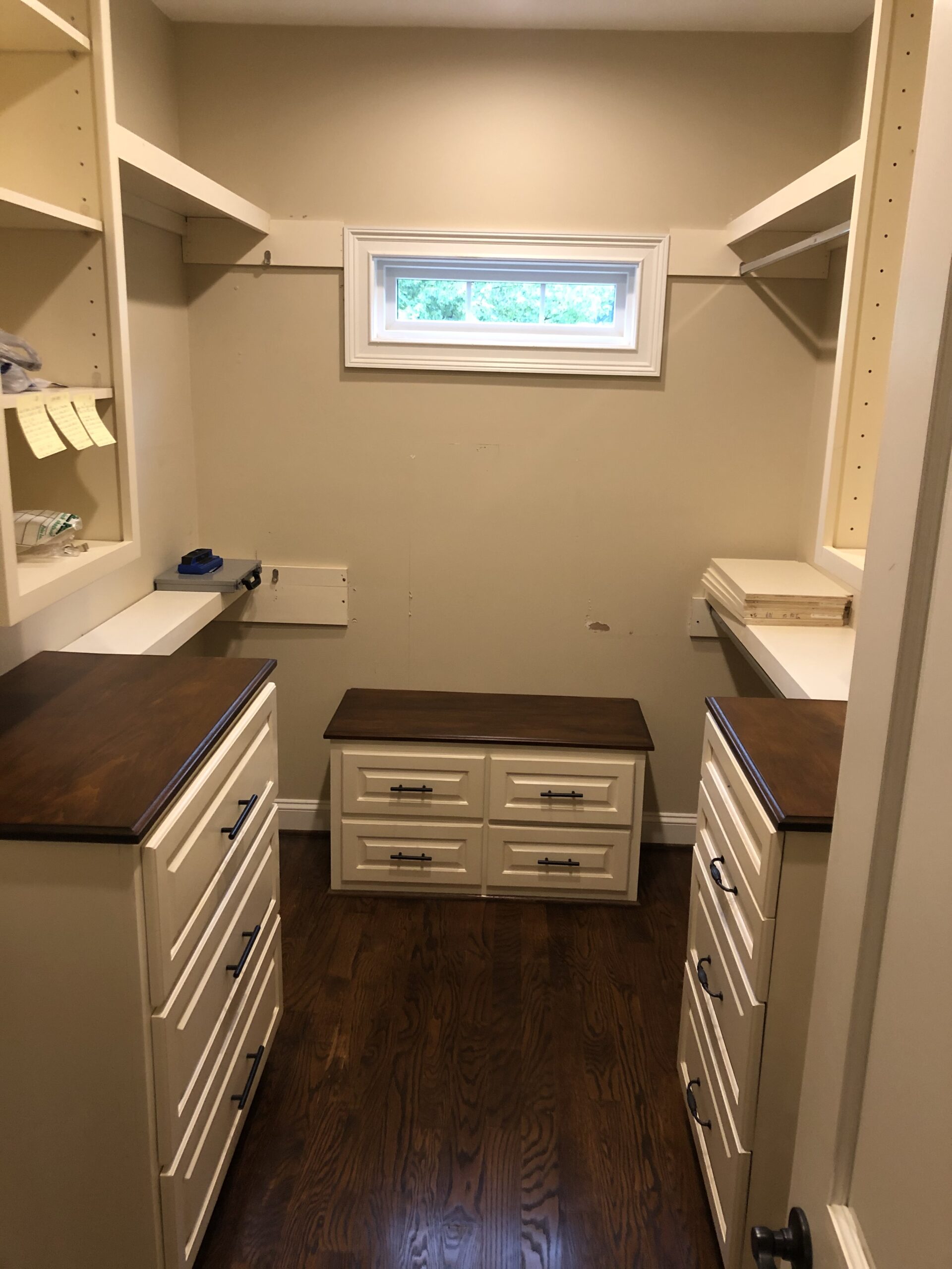 A walk-in closet with wooden floors, built-in shelves, drawers, and a small window at the top.