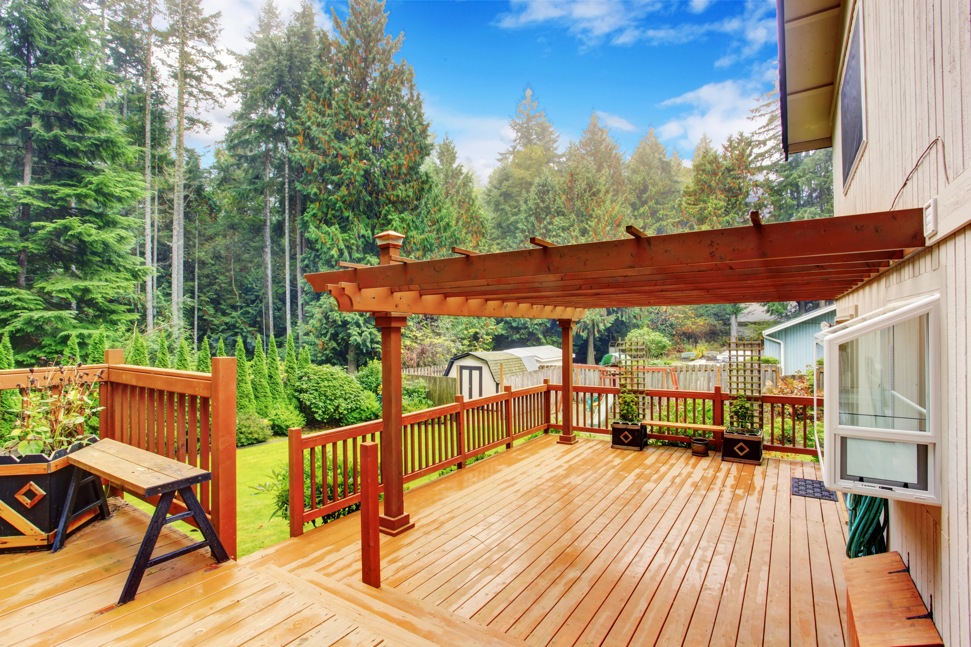 A wooden deck with a pergola overlooks a green backyard bordered by tall trees. A small shed is visible in the background.