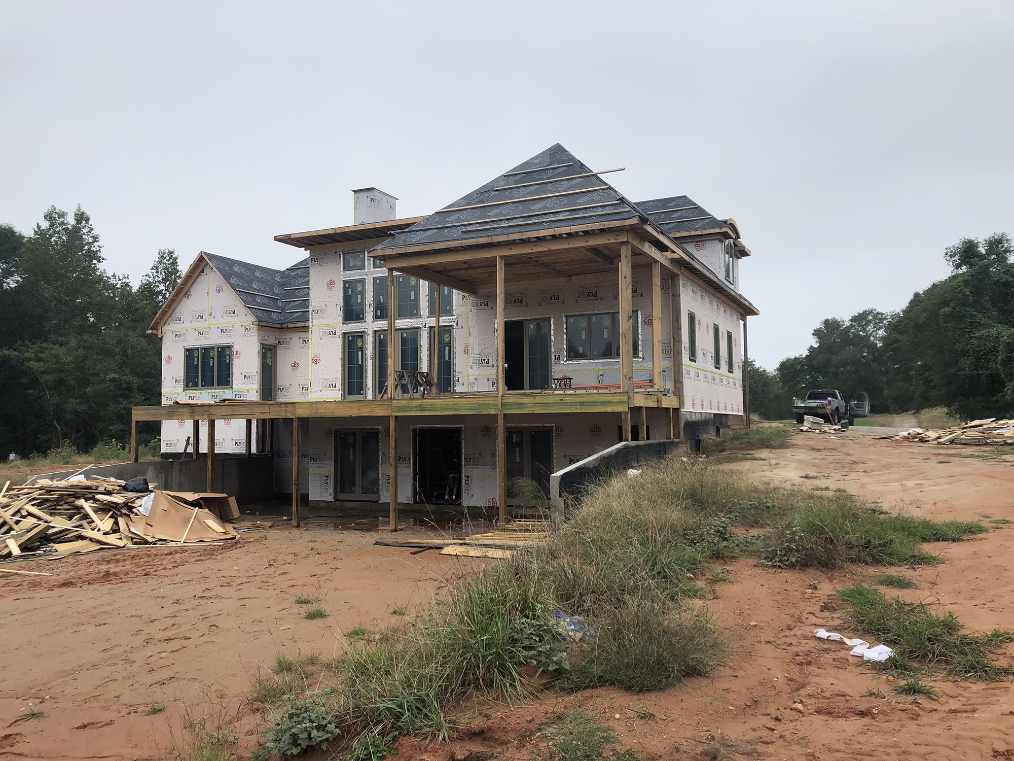 Two-story house under construction with exposed wooden framing, windows installed, and roofing partially completed. Surrounding area shows construction debris and unfinished landscaping.