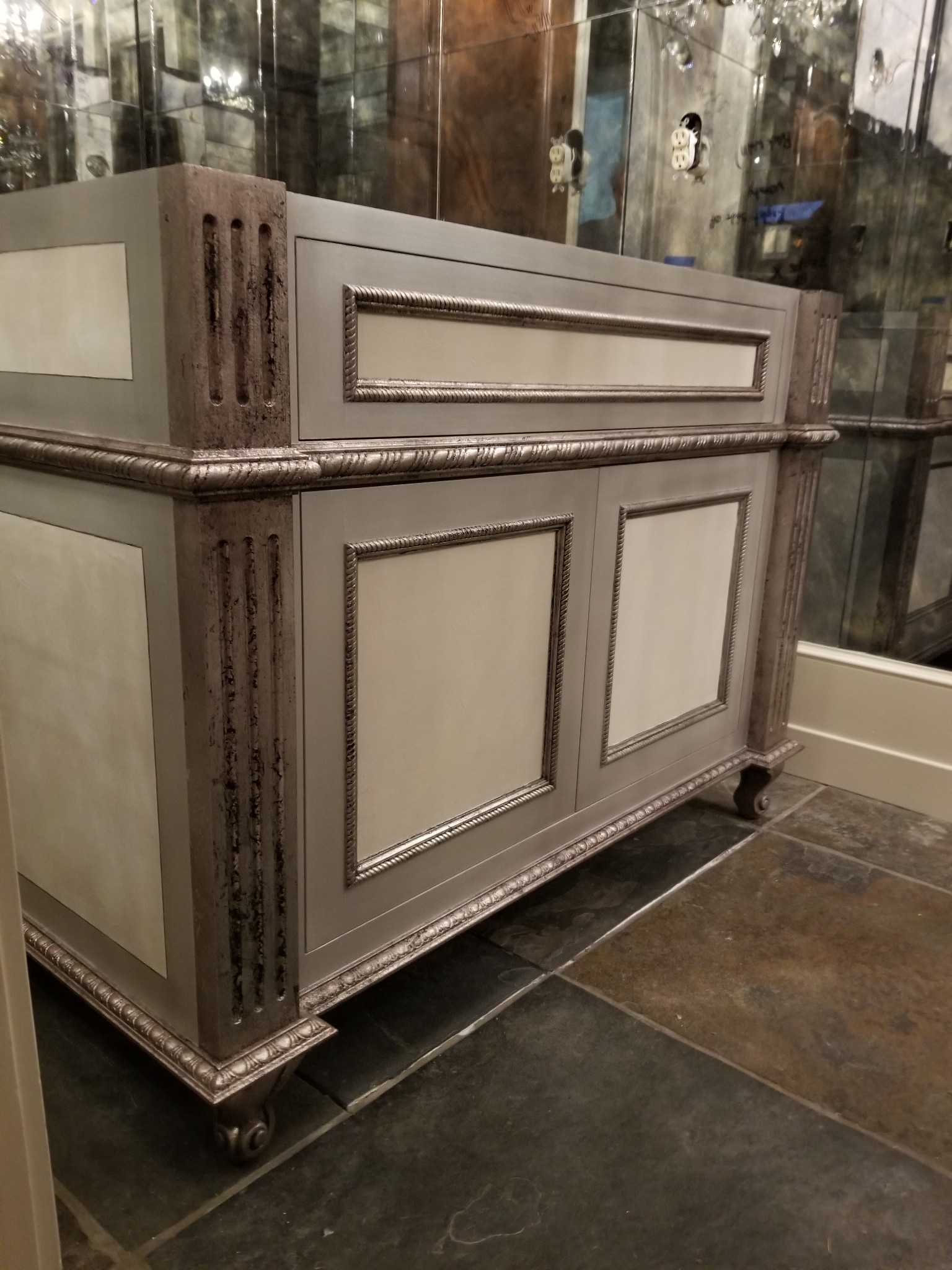 Elegant, vintage-style vanity cabinet with decorative panels and metallic accents, set on a stone-tiled floor in a mirrored room.