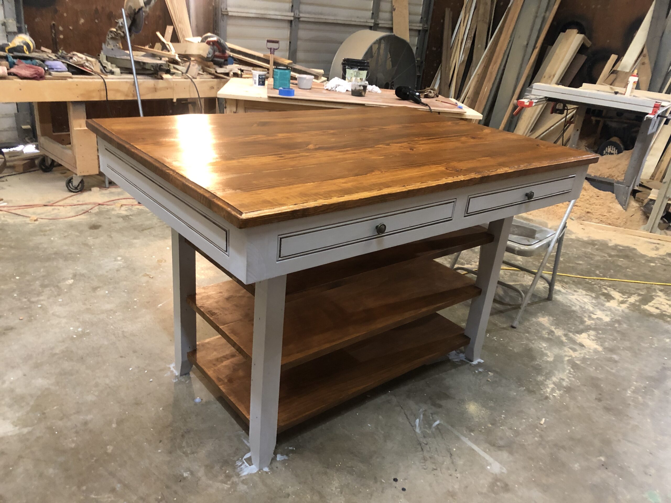 A wooden workshop table with a polished top, two drawers, and two shelves in a garage workspace. Tools and materials are visible in the background.