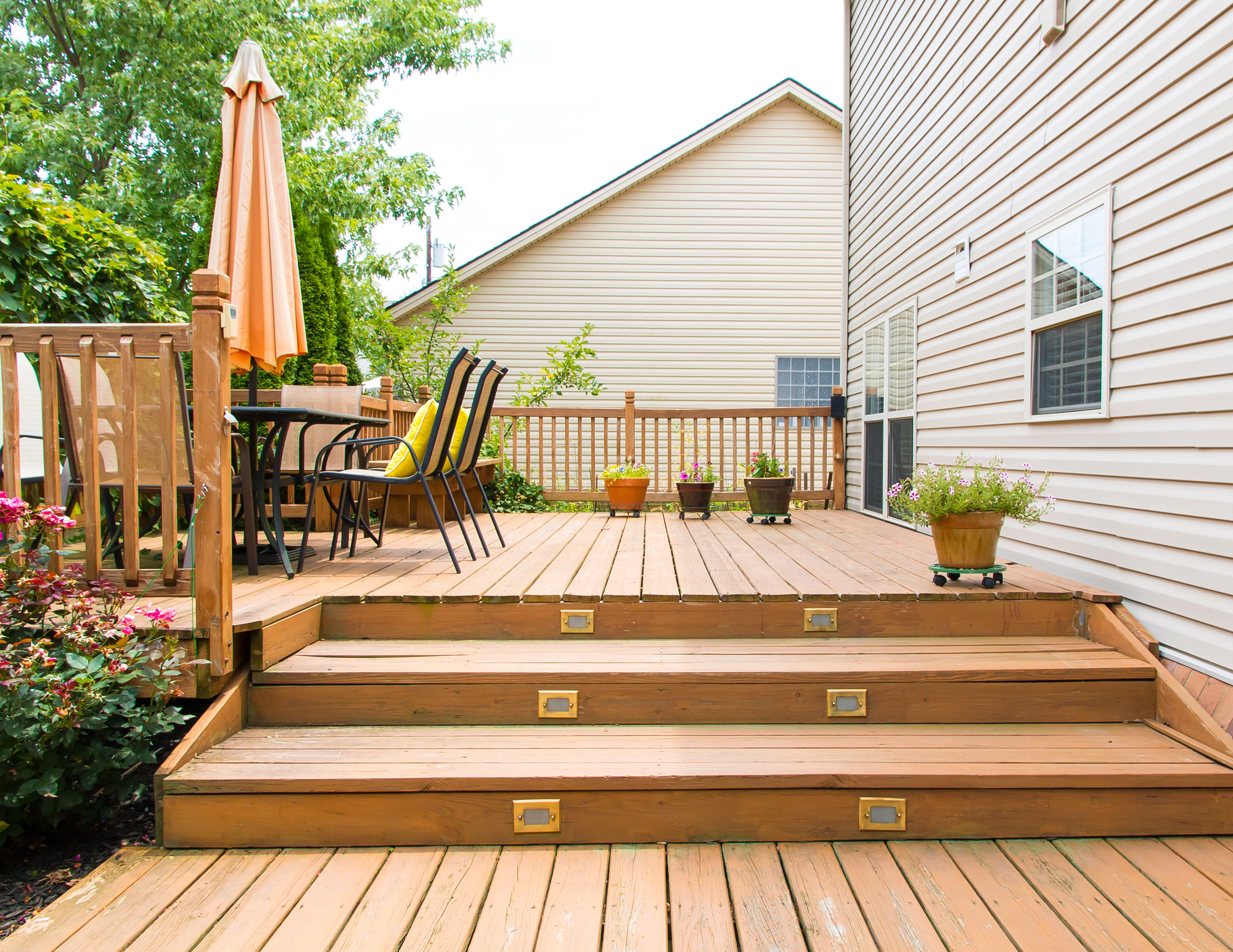 A wooden deck with steps features a patio table with an umbrella, surrounded by chairs. Potted plants decorate the space, and a house and trees are visible in the background.