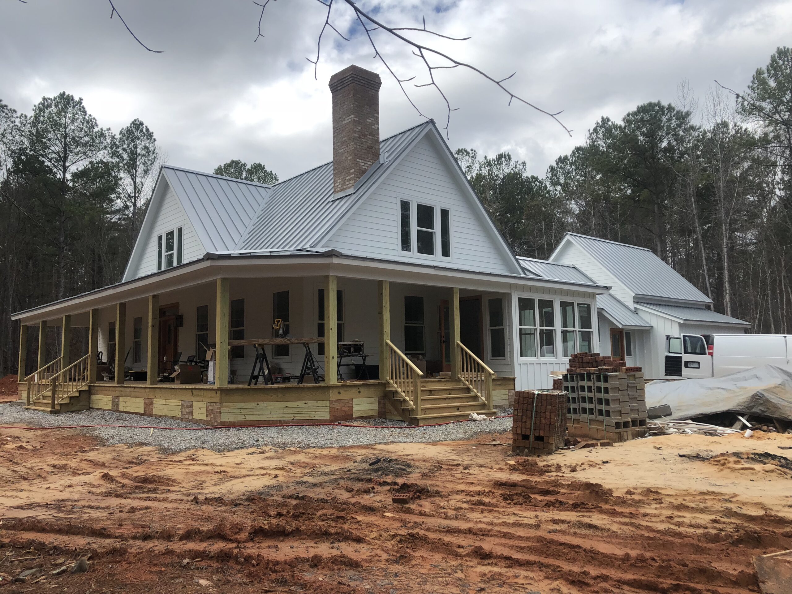 A white house under construction with a wrap-around porch and brick chimney, surrounded by trees and piles of construction materials.