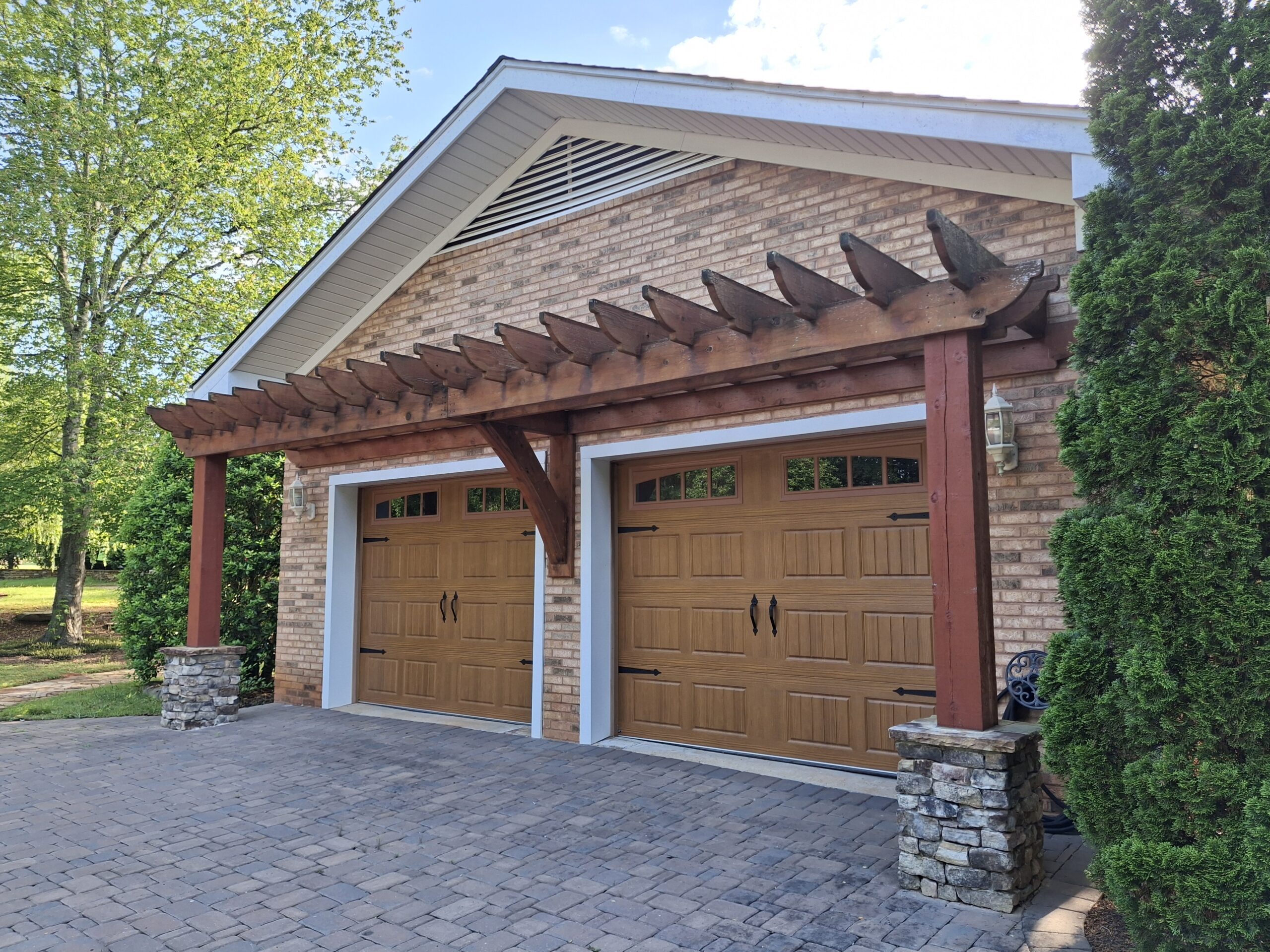 A brick garage with two wooden doors and a wooden pergola overhead, set on a paved driveway, surrounded by trees and greenery.