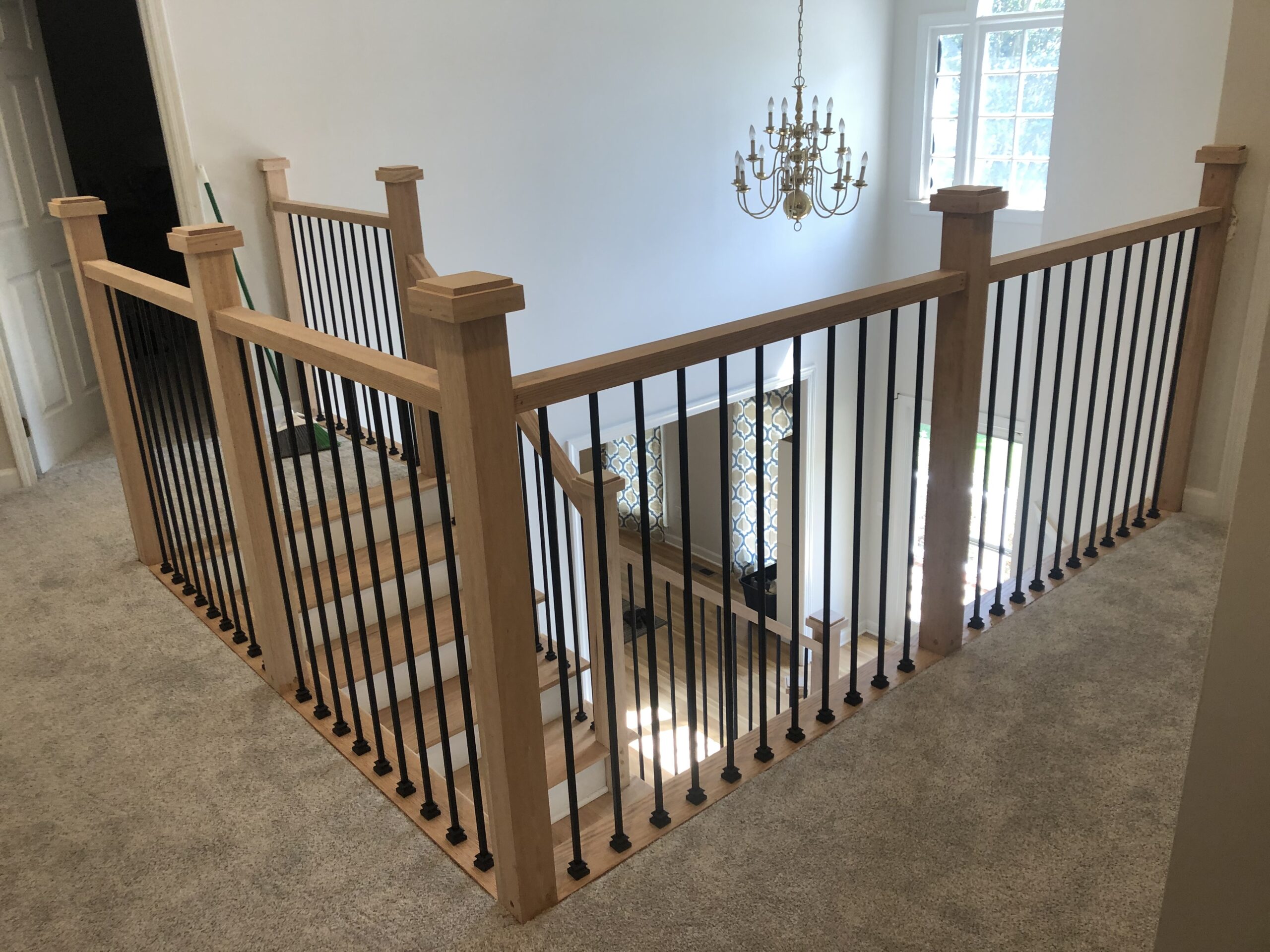 Wooden staircase with black metal balusters and carpeted flooring, featuring a chandelier above and a window in the background.