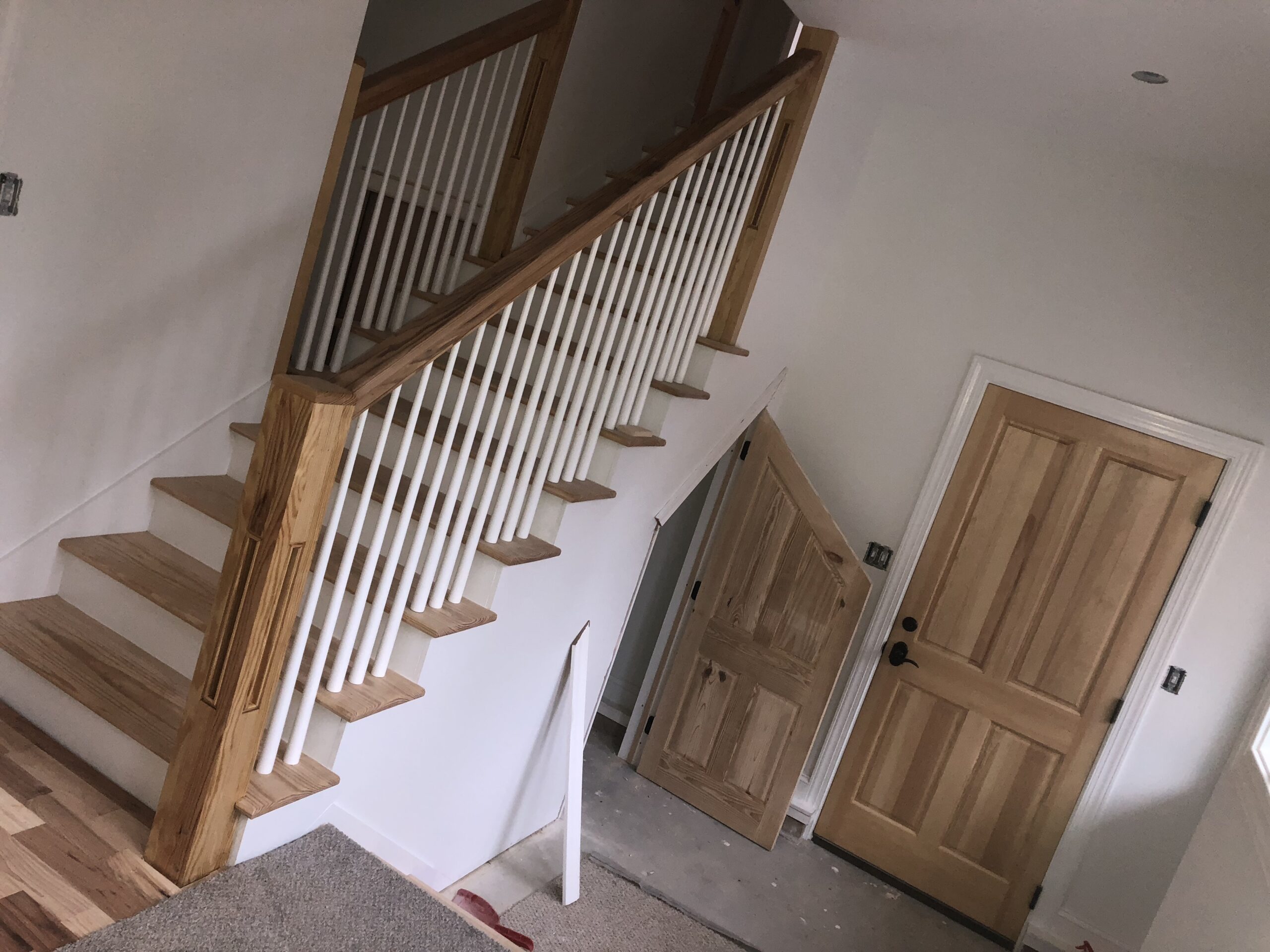 Stairway with wooden handrails and white balusters, partially finished with carpet. A wooden door stands ajar on the right. Construction or remodeling is ongoing.