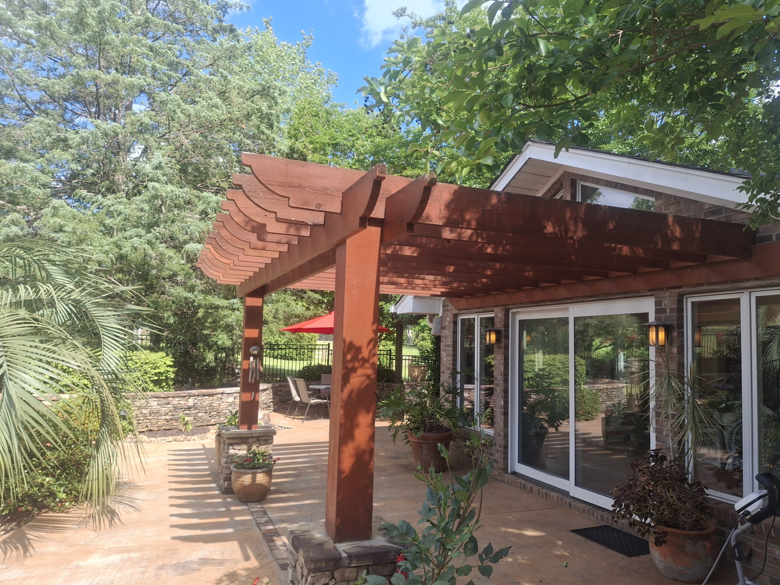A wooden pergola extends over a patio next to a house with large glass doors, surrounded by potted plants and greenery under a blue sky.