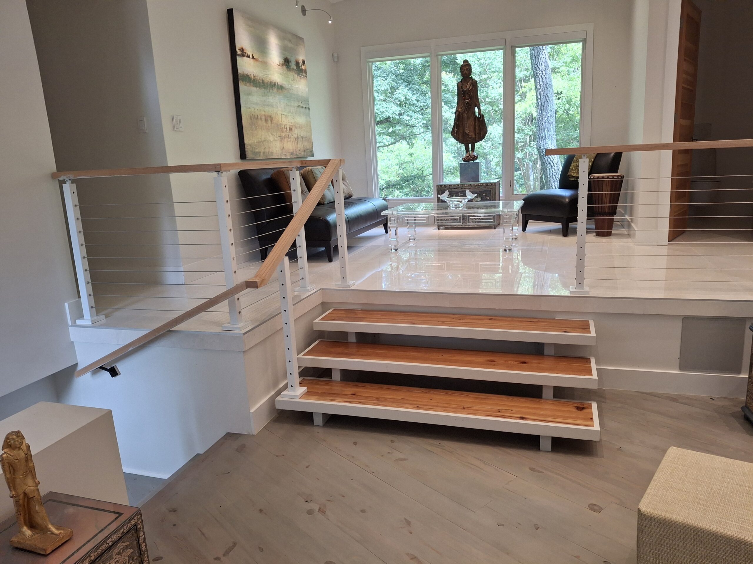 A modern living area with a small elevated section, featuring two chairs, a coffee table, a statue, and large windows overlooking trees. Steps with wooden treads lead to the elevated space.