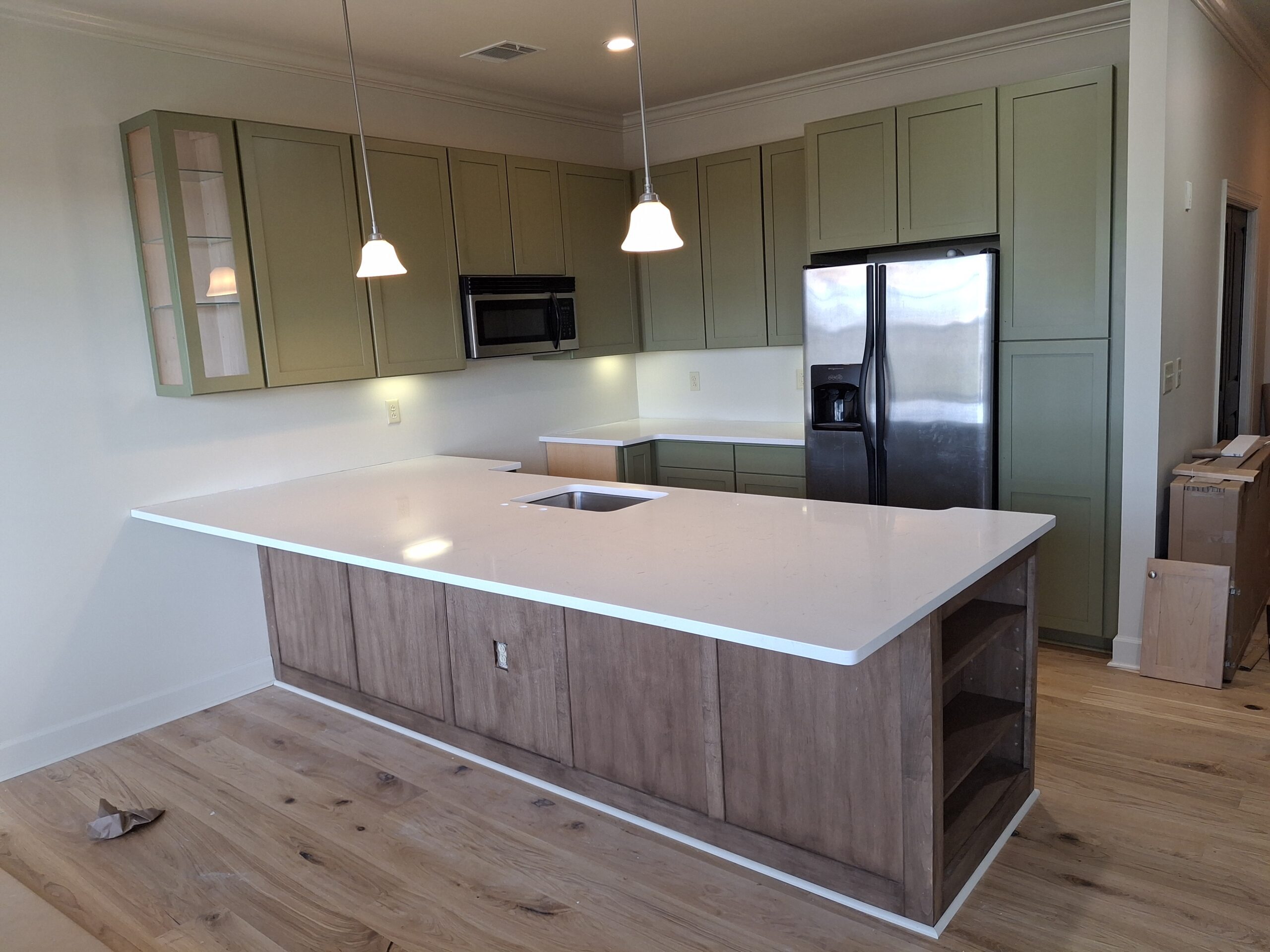 Modern kitchen with green cabinets, stainless steel appliances, a white island countertop with sink, pendant lights, and light wood flooring.