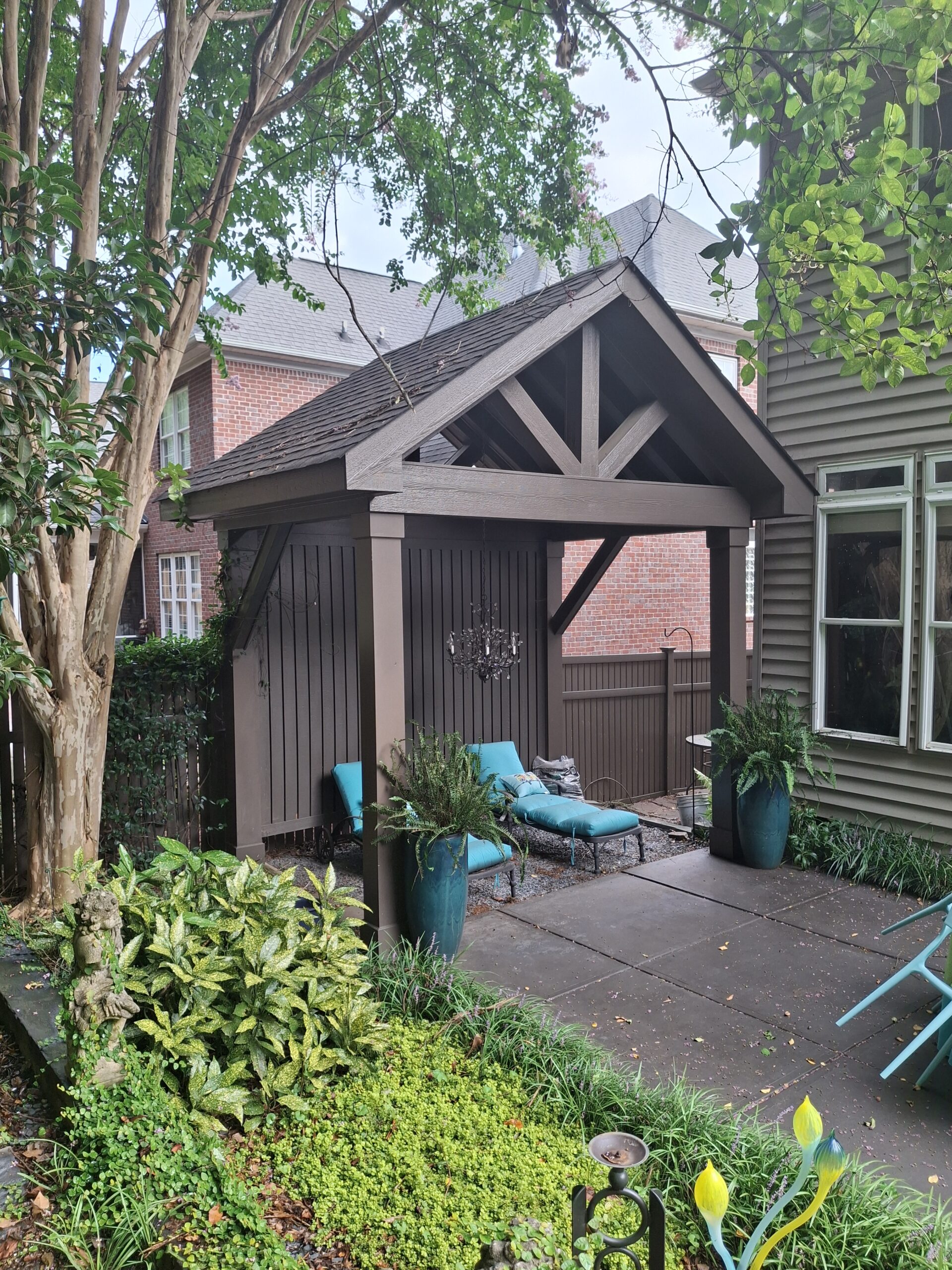 A small wooden gazebo with a pitched roof is in a garden. It contains a blue lounge chair and a small chandelier. Surrounding greenery includes a tree and potted plants.